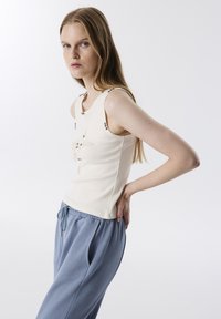 Getailleerde ivoorkleurige tanktop met decoratieve zilveren knopen op de schouderbanden, voorzien van bloemapplicaties aan de voorkant, gecombineerd met lichtblauwe sweatpants.