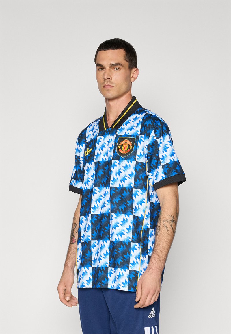 Blau-schwarz karierter kurzärmeliger Jersey mit Kragen, der ein Manchester United-Wappen und gelbe Akzente aufweist. Der Stoff wirkt leicht.