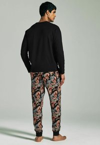 Schwarzes Langarmoberteil kombiniert mit gemusterten Hosen, die rote, blaue und cremefarbene Formen auf einem schwarzen Hintergrund zeigen; elastische Bündchen sichtbar.