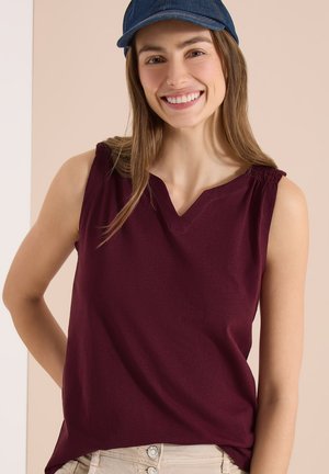 Femme souriante portant un haut sans manches rouge foncé, un pantalon beige et une casquette en jean bleue sur un fond beige.