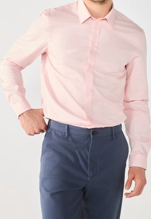 Mann trägt ein hellrosa langärmliges Hemd, das in dunkelblaue Hose gesteckt ist, mit einer Hand in der Tasche vor einfarbigem Hintergrund.