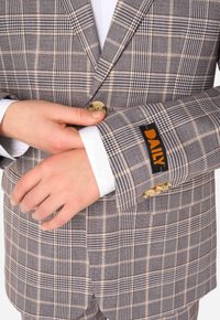 Blazer a quadri marrone con un motivo a righe leggere, con bottoni dorati e un'etichetta arancione "DAILY" sul polsino, tenuto da mani di carnagione chiara.