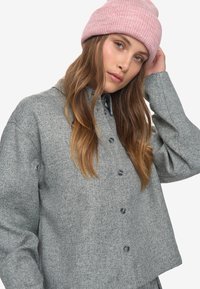 Junge Frau mit langen braunen Haaren, die ein graues Knopfhemd und eine rosafarbene Strickmütze trägt und mit einer Hand nahe ihrem Kopf posiert.