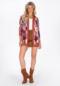Donna che indossa un cardigan rustico a fiori, una maglietta bianca, una mini gonna in suede marrone e stivaletti alla caviglia in suede marrone abbinati, in piedi contro uno sfondo bianco.