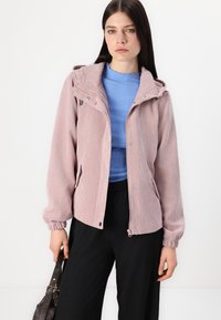 Rosa kordfløyelsjakke med hette, frontglidelås, trykknapper og sidelommer. Brukt over en blå ribbet genser og svarte bukser.
