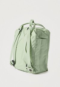 Zaļš auduma mugursoma ar regulējamām siksnām, kvadrāta formas, sānu kabatu un gludu tekstūru. Redzami sprādzes un šuves.
