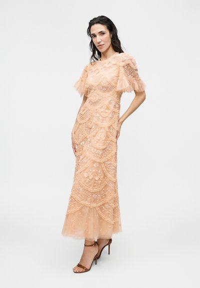 Γυναίκα που στέκεται φορώντας μακρύ μέχρι το πάτωμα ροζ δαντελένιο φόρεμα με φουσκωτά μανίκια και καφέ πέδιλα με ψηλό τακούνι, σε απλό φόντο.