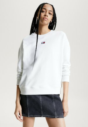 Felpa bianca con polsini e orlo a costine, decorata con un piccolo logo rettangolare. Abbinata a una mini gonna in denim scuro con cuciture verticali.