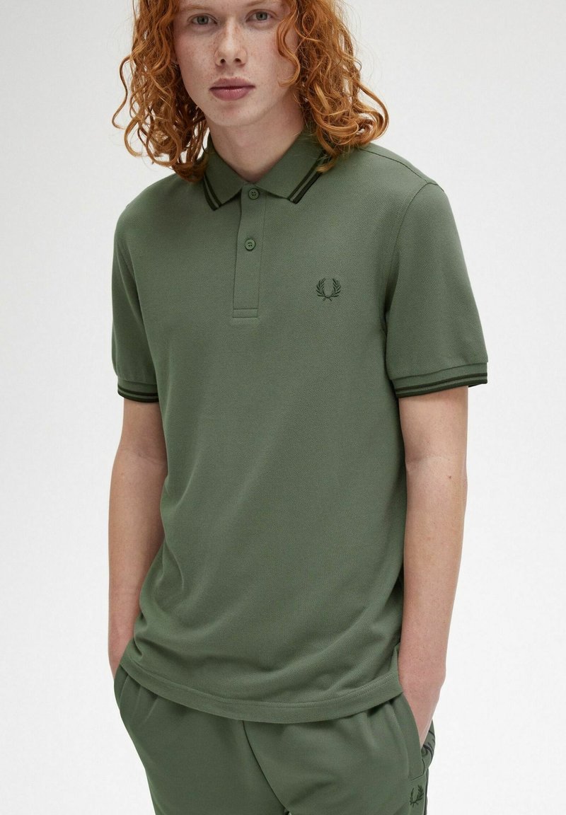 Camisa polo verde con tejido texturizado, tapeta de tres botones y acentos en negro en el cuello y las mangas. Pequeño logo en el pecho.