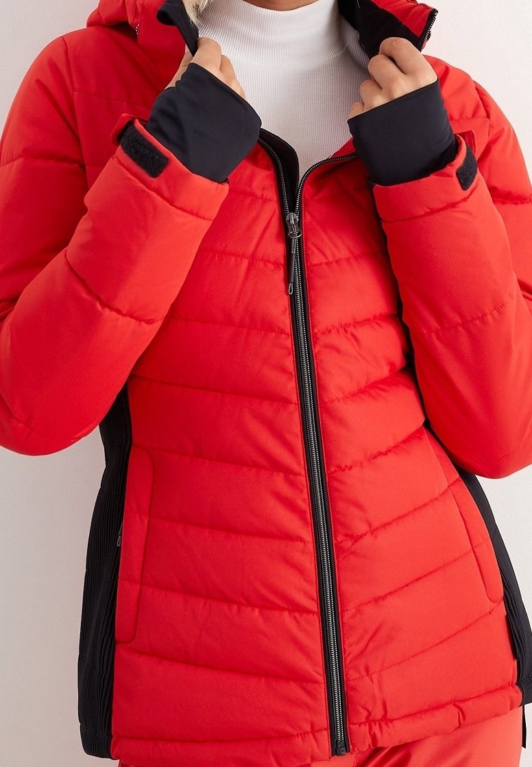 Personne portant une veste d’hiver matelassée rouge avec des panneaux latéraux et des poignets noirs, fermant ou ajustant le col avant sur un col roulé blanc.