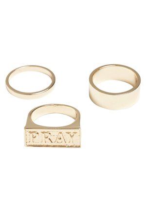 Tre anelli d'oro: un anello sottile, un anello largo e liscio, e un anello rettangolare inciso con la parola "PRAY" in una finitura texturizzata.