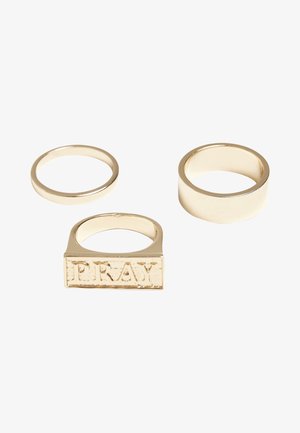 Tri zlate prstane: en tanek obroč, en širok gladek obroč in en pravokotni obroč z vgraviranim napisom "PRAY" v teksturiranem finišu.