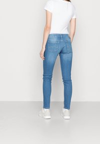 Kvinna i ljusblå skinny jeans och vita sneakers, står med ryggen mot kameran mot en enkel vit bakgrund.