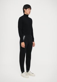 Jovem vestido com um suéter de gola alta preto, calças de treino pretas com bolsos com fecho, meias escuras e tênis brancos, posando contra um fundo liso.