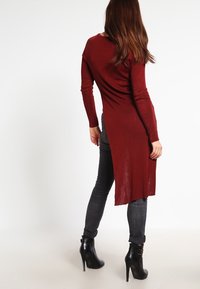 Langer bordeauxfarbener Cardigan mit geradem Schnitt und asymmetrischem Saum, aus leichtem Stoff, kombiniert mit dunklen Jeans und schwarzen Stöckelschuhen.