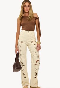 Top marrón de un solo hombro combinado con jeans beige de talle alto que presentan recortes florales, bolsillos planos y un diseño relajado y acampanado.