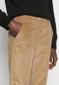 Pantalones de corduroy marrón claro con textura acanalada, bolsillos laterales y corte recto, combinados con un suéter negro.