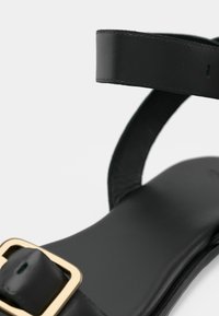 Μαύρο δερμάτινο σανδάλι με επίπεδη σόλα, έναν ιμάντα και χρυσή αγκράφα. Λεπτό υφή και μινιμαλιστικός σχεδιασμός.