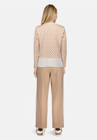 Veste beige à motifs avec manches longues, associée à un pantalon beige. La veste présente un design géométrique et un aspect superposé, avec une texture lisse.