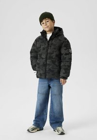 Doudoune camouflage noire avec fermeture éclair à l'avant, grandes poches et capuche. Associée à un jean en denim bleu ample et des baskets aux tons neutres.