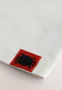 Tissu blanc avec un patch carré rouge présentant un motif d’araignée noire. Le patch a une finition texturée avec un détail de toile d’araignée.