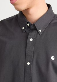 Camicia grigia scura con bottoni, colletto appuntito, bottoni bianchi in madreperla e piccolo logo bianco sul petto a sinistra. Materiale in cotone.