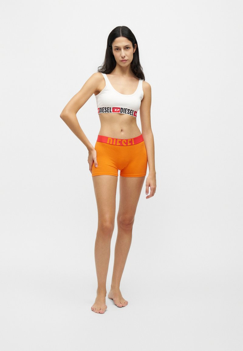 Femme portant un soutien-gorge de sport blanc et un short boxer orange avec des bandes rouges à la taille, tous deux estampillés du mot Diesel, debout pieds nus sur fond blanc.
