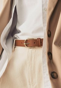 Brown leather belt with a gold metal buckle, worn over cream-colored pants and concealed under a light tan jacket and white shirt.
