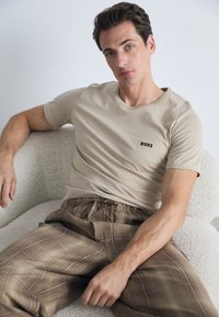 Beige cotton t-shirt with a black logo on the chest, paired with brown checkered pants featuring an elastic waistband. Textured background.