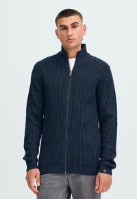 Mørk marineblå strikket glidelås cardigan med ribbestruktur, høyt krage og lange ermer. Har glidelås foran og logo-patch på mansjetten.