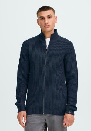 Mørk marineblå strikket glidelås cardigan med ribbestruktur, høyt krage og lange ermer. Har glidelås foran og logo-patch på mansjetten.
