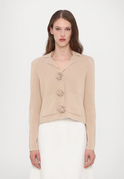 Beige cropped jas gemaakt van textuurstof, met drie bloemvormige knopen en twee voorzakken met een notch-kraagontwerp.