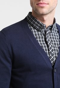 Marinblå stickad cardigan med V-ringning, lager på lager över en svart-och-vit rutig skjorta, med knappdetaljer och en mjuk textur.