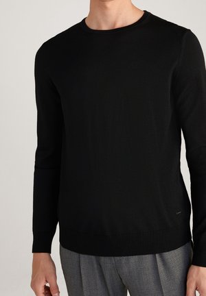 Mann trägt einen schlichten schwarzen Langarmpullover und graue karierte Hose, steht vor einem schlichten hellen Hintergrund.