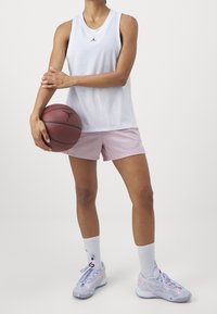 Vit ärmlös linne med svart logotyp, rosa meshshorts och flerfärgade sneakers, håller en basketboll.