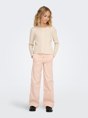 Béžový rebrovaný kardigán s okrúhlym výstrihom a zapínaním na gombíky, v kombinácii so svetloružovými nohavicami so širokými nohavicami a čiernymi topánkami, ktoré nosí dieťa.