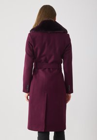 Paarse wollen jas met een bontkraag, een ceintuur in de taille, lange mouwen en knoopaccenten op de manchet. Ontworpen met een rechte achterkant en zijslits.