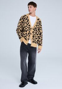 Cardigan a stampa leopardata in beige e nero, con scollatura a V, chiusura con bottoni e due tasche anteriori, abbinato a jeans grigio scuro.