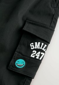 Un pantalon cargo noir est doté d'une poche latérale avec une impression blanche "SMIL 247" et un patch rond turquoise avec un détail de visage souriant.