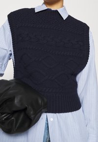 Person wearing navy blue cable-knit sleeveless sweater over light blue striped shirt, holding a black leather clutch bag under arm.