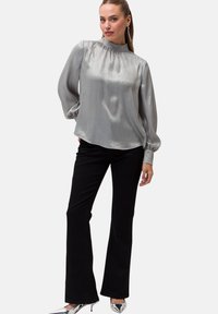 Blusa argento a maniche lunghe con collo alto, texture morbida e dettagli arricciati sulle spalle, abbinata a pantaloni svasati neri.