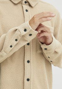 Beige fleece skjorte med teksturert overflate, knappelukking foran, svarte knapper og krage. Har rullede mansjetter og en enkelt brystlomme.