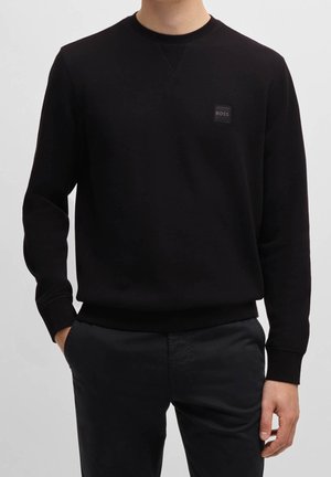 Homme portant un sweat-shirt noir à col rond à manches longues avec un petit logo "BOSS" sur la poitrine et un pantalon noir, debout les mains dans les poches.
