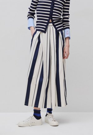 Frau trägt marineblaue und weiß gestreifte Palazzohosen, marineblauen gestreiften Cardigan, passende Socken und weiße Sneakers, steht mit den Händen in den Taschen.