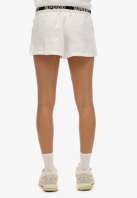 Pantaloni corti bianchi in lino con vita elasticizzata e il marchio "SUPERDRY". Indossati con scarpe chiare e calzini bianchi, mostrano una vestibilità rilassata.