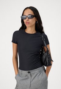 Black fitted short-sleeve top paired with gray tailored trousers; model holds a small black leather bag and wears rectangular sunglasses.
