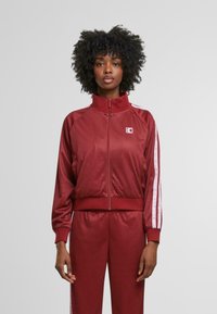 Červená sportovní bunda s vpředu na zip, vysokým límcem a bílými pruhy na rukávech. Vyrobena z prodyšného materiálu s texturovaným vzorem.