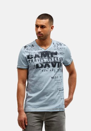 Männliches Model trägt ein hellblaues T-Shirt mit V-Ausschnitt und Grafik sowie graue Hose, steht mit einer Hand in der Tasche vor einfachem Hintergrund.