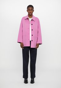 Pink wool coat with a collar, front buttons, and two side pockets, paired with dark pinstriped trousers and black ankle boots.