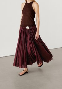Haut marron sans manches associé à une jupe maxi fluide bordeaux avec des plis et une ceinture décorative. Des sandales noires à lanières complètent la tenue.